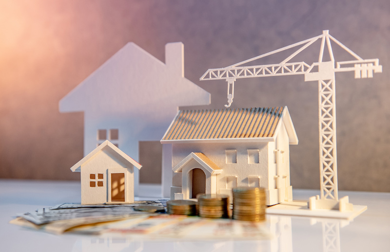 Coin stack on international banknotes with house and construction crane models on the table.