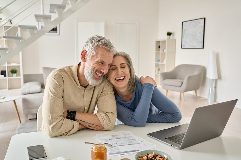 Happy mature older family couple laughing, bonding sitting at home table with laptop.